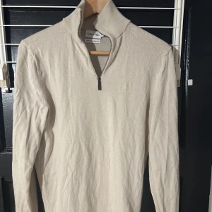 Beige tröja med dragkedja från Calvin Klein - Säljer en stilren beige tröja från Calvin Klein i maskintvättbar ull. Tröjan har en halv dragkedja och en diskret CK-logga på bröstet. Perfekt för en elegant och avslappnad look.
