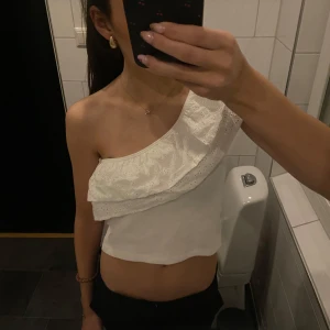 Vit oneshoulder topp med volang - Säljer en söt vit oneshoulder topp med volangdetaljer. Perfekt för sommarens varma dagar. Toppen har en fin broderad design och är tillverkad i ett lätt och bekvämt material. Helt ny har prislappen kvar, aldrig använd💕
