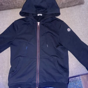 Moncler kofta - Säljer en snygg marinblå kofta från Moncler med dragkedja och huva. Den är i nyskick och den har inga skador. Har haft den i 1 och ett halvt år men har ej använt den så mycket eftersom jag inte gillade hur den satt på mig. Den är storlek S. Den har inga skador eller liknande. Vid snabb affär kan sänka priset! Skriv till mig om fler funderingar eller tankar. Mvh Alex