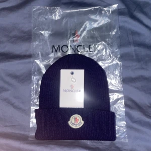 Moncler mössa äkta - Snygg svart ribbad mössa från Moncler med broderad logotyp framtill. Perfekt för att hålla värmen med stil. Mössan är i ett klassiskt utförande och kommer med originalförpackning och etikett.