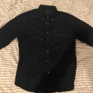 Mörkblå manchester skjorta från East West - Snygg mörkblå manchester skjorta från East West med klassisk krage och knappar framtill. Perfekt för en stilren look. Skjortan har långa ärmar och är tillverkad i mjukt material för extra komfort.