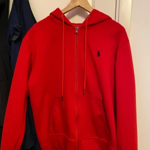 Röd hoodie från Ralph Lauren - Säljer en snygg röd hoodie från Ralph Lauren med dragkedja och klassisk logga på bröstet. Den har en justerbar huva med snören och praktiska fickor framtill. Perfekt för en avslappnad stil.