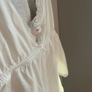 Vit linne med spetsdetaljer - Säljer ett vitt linne från Nelly med spetsdetaljer längs halsringningen. Linnet har smala axelband och en v-ringad design. 