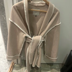 Beige Frenchie wool kappa från Neo Noir - Säljer en elegant beige kappa från Neo Noir. Den har en stilren design med vita detaljer längs kanterna och ett knytbälte i midjan. Tillverkad i en blandning av ull och polyester för en mjuk och bekväm känsla. Perfekt för att hålla stilen under kyligare dagar. Perfekt nu till våren och sommaren! Storlek 36