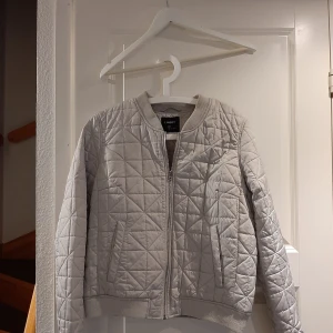 Grå quiltad jacka från Lindex - Säljer en stilren grå quiltad jacka från Lindex. Jackan har en dragkedja framtill och ribbade muddar vid ärmslut och nederkant. Perfekt för kyligare dagar med sin bekväma passform och snygga design.