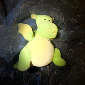 Gosedjur i form av en drake - Söt och mjuk drake i ljusgrönt och gult tyg. Den har små vingar och en taggig rygg i grått. Perfekt som kramkompis eller dekoration i barnrummet.