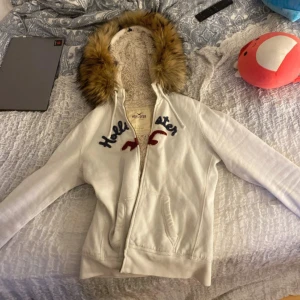 Zip up fur hoodie med päls från Hollister - Säljer en vit hoodie från Hollister med äkta pälsfodrad luva. Pälskragen är EGENSYDD! Storlek L men passar S bäst, skriv gärna innan ni köper, priset går att diskuteras