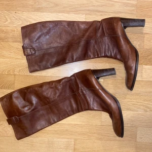 Bruna skinnstövlar från Rizzo 40,5 - Säljer ett par bruna skinnstövlar med klack. De har en stilren design och är perfekta för höst och vinter. Stövlarna har en dekorativ remdetalj upptill och en robust sula.