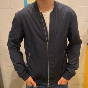 Mörkblå bomberjacka från J.Lindeberg - Säljer en stilren mörkblå bomberjacka från J.Lindeberg. Jackan har dragkedja framtill och två fickor med dragkedjor. Den är långärmad och har en ribbad krage samt nederkant. Perfekt för en cool och avslappnad look. Liten repa höger om vänster ficka ingenting som stör. Annars fint skick, modellen är 182 och 72 kg passar. Nypris 1899 kr, unik modell. Pris kan diskuteras