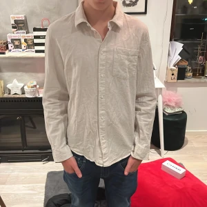 Beige långärmad linne skjorta från Lindex  - Säljer en stilren beige långärmad skjorta med klassisk krage och bröstficka. Perfekt för en avslappnad men ändå snygg look. Skjortan har en normal passform och är tillverkad i linne. Skick 10/10 aldrig använt.