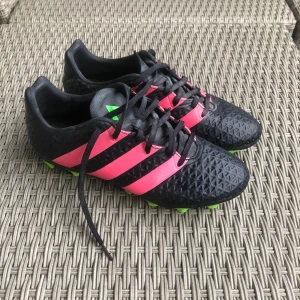 Svarta och rosa fotbollsskor från Adidas - Snygga fotbollsskor från Adidas i svart och rosa med gröna dobbar. Säljes pga av att de är för små