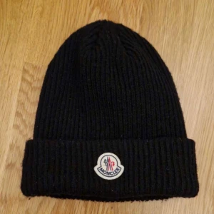 Svart Moncler Mössa - Snygg svart ribbad mössa från Moncler med uppvikt kant och logotyp framtill. Perfekt för kyliga dagar och ger en stilren look. Den är i fint skick utan några skador alls. Den har använts lite men inget som märks. Nypris ligger på cirka 2999kr medans mitt pris är 599kr. Kan ej garantera autenticitet då den var en present som jag fick för ett tag sen, men kvaliten är riktigt bra. Hör gärna av er om ni har några frågor/bud! 