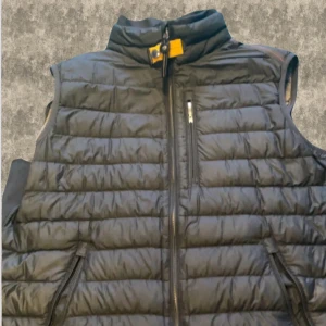 Svart dunväst från Parajumpers - Snygg svart dunväst från Parajumpers med dragkedja och hög krage. Västen har en praktisk bröstficka med dragkedja och märkets logotyp på insidan. Perfekt för kyliga dagar. Några mindre defekter