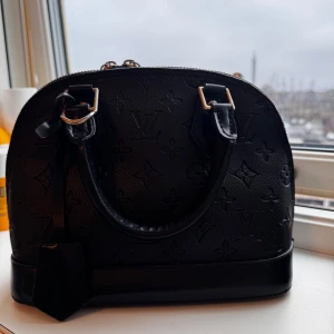 Svart handväska med monogram - Elegant svart handväska i skinn med monogrammönster och guldfärgade detaljer. Väskan har dubbla handtag och en dragkedja upptill. Perfekt för att ge en lyxig touch till din outfit.