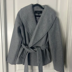 Grå kappa från Vero Moda - Snygg grå kappa från Vero Moda med knytbälte i midjan. Den har en elegant omlott-design och långa ärmar. Perfekt för kyligare dagar.