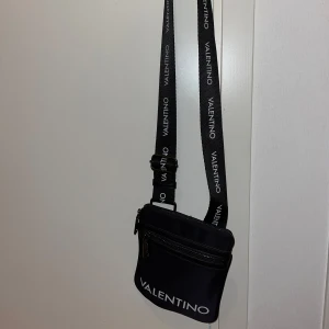 Svart axelväska från Valentino - Snygg svart axelväska från Valentino med justerbar axelrem och dragkedja. Väskan har en stilren design med logotypen tryckt i vitt på både remmen och framsidan. Perfekt för att bära med sig det nödvändigaste med stil. Är. 4 fickor oså iafall jätte fint skick knappt använd