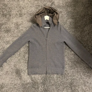 Grå ulltröja med huva från Massimo Dutti - Snygg grå ulltröja från Massimo Dutti i storlek M med avtagbar brun huva.  ribbstickad för extra stil. Perfekt för kyliga dagar. Cond 8/10