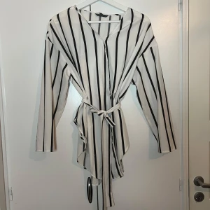 Vit och svart randig klänning från shein - Snygg vit och svart randig klänning från shein med knytband i midjan. Klänningen har långa ärmar och en avslappnad passform. Perfekt för en stilren look.