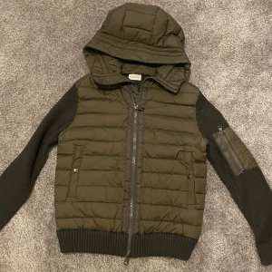 Grön/mörkgrön jacka från Moncler - Säljer en snygg olivgrön jacka från Moncler i mycket bra skick. Jackan har en quiltad design med dragkedja och huva. Ärmarna är långärmade och stickade, vilket ger en cool kontrast. Perfekt för höst och vår!