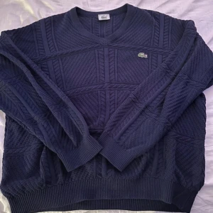 Mörkblå stickad tröja från Lacoste - Säljer en snygg mörkblå stickad tröja från Lacoste med det klassiska krokodilmärket på bröstet. Tröjan har en v-ringad hals och ribbade muddar vid ärmslut och nederkant. Perfekt för kyliga dagar!