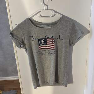 Säljer en grå t-shirt från Bondelid i storlek XS. Den har ett tryck med amerikansk flagga och texten 'Genuine Design - Finest Quality'. Perfekt för en avslappnad stil. T-shirten är kortärmad och i mycket bra skick. Knappast använd.