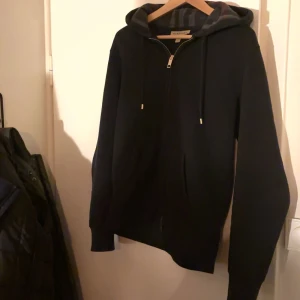 Svart hoodie från Burberry - Säljer en stilren svart äkta hoodie från Burberry i storlek M. Den har en dragkedja framtill och en broderad logga på bröstet. Perfekt för höst och vår, med en bekväm passform och långa ärmar. Huvan har justerbara snören för extra komfort. Passar både till vardags och lite finare tillfällen. 