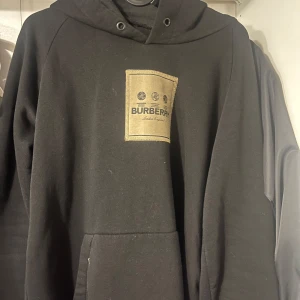 Svart hoodie från Burberry - Snygg svart hoodie från Burberry med en beige patch på bröstet. Hoodien har en klassisk design med känguruficka och justerbar huva. Perfekt för en stilren och bekväm look.