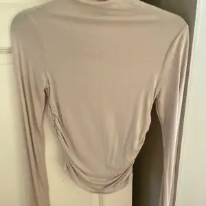 Säljer en stilren beige långärmad topp i mjukt material. Toppen har en elegant rynkad detalj på sidan som ger en fin passform. Perfekt för både vardag och festliga tillfällen. Passar bra till jeans eller kjol för en chic look.