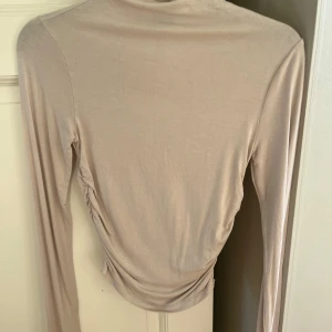 Beige långärmad topp - Säljer en stilren beige långärmad topp i mjukt material. Toppen har en elegant rynkad detalj på sidan som ger en fin passform. Perfekt för både vardag och festliga tillfällen. Passar bra till jeans eller kjol för en chic look.