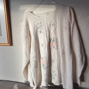 Vit stickad tröja med blommönster - Vit stickad tröja med ett subtilt blommönster i rosa och grönt. Tröjan har långa ärmar och en rund halsringning, perfekt för kyliga dagar. Den har en klassisk och romantisk stil som passar bra till både jeans och kjol.