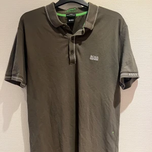 kortärmad pikétröja från Hugo Boss - Snygg pikétröja från Hugo Boss med korta ärmar och vit logga på bröstet. Tröjan har en klassisk krage med knappar och kontrasterande detaljer i grönt. Perfekt för en stilren look.