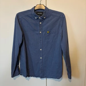 Blå skjorta från Lyle & Scott - Snygg blå skjorta från Lyle & Scott med klassisk krage och knappstängning. Skjortan har en bröstficka med det ikoniska Lyle & Scott-emblemet. Perfekt för en stilren look.