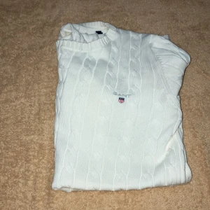 Vit stickad tröja med broderad logga - Säljer en snygg vit stickad tröja i storlek S. Den har ett klassiskt flätmönster och en liten gant  logga på bröstet. Perfekt för både höst och vinter, och den är supermjuk och bekväm. Passar till alla tillfällen, från skolan till en mysig fika. 🧶✨