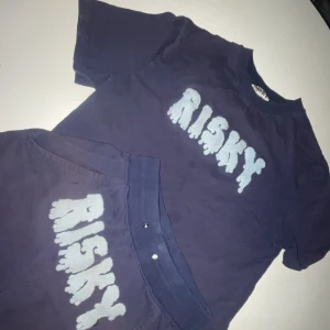 Mörkblå t-shirt/shorts från Risky - Cool mörkblå t-shirt från Risky med ett stort, vitt tryck på bröstet. Perfekt för en avslappnad stil och passar bra till både jeans och shorts. T-shirten är kortärmad och gjord i ett mjukt material för extra komfort.