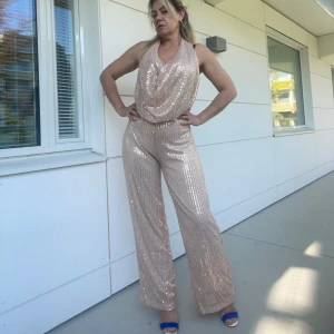 Glittrig jumpsuit i guld - Snygg glittrig jumpsuit i guld med vida ben och djup urringning. Perfekt för en festlig kväll med sitt iögonfallande paljettmönster. Jumpsuiten har en lös passform och är ärmlös, vilket ger en elegant och bekväm stil.helt ny