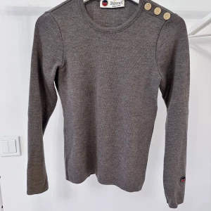 Grå tröja med knappar från Busnel - Snygg grå tröja från Busnel med dekorativa knappar på axeln och ärmslut. Tröjan har en rund halsringning och är tillverkad i 100% merinoull.