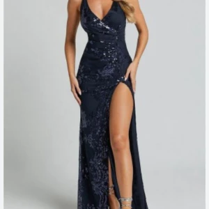 Blå glittrig långklänning - Elegant blå långklänning med glittriga detaljer och djup slits. Klänningen har en v-ringad design och smala axelband som korsas i ryggen, vilket ger en stilfull look.