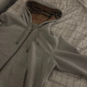  Burberry kofta - Säljer en stilren grå hoodie från Burberry i 100% bomull. Den har en klassisk dragkedja och en fodrad huva med det ikoniska Burberry-mönstret. Perfekt för en avslappnad och trendig look koftan har använts lite fast det har inga skador och är typ helt ny för mer information kontakta mig 