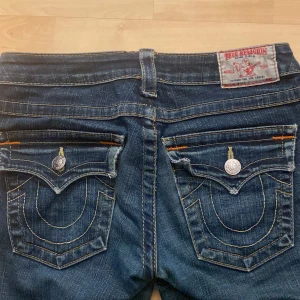 Mörkblå jeans från True Religion - Snygga mörkblå jeans från True Religion med ikoniska bakfickor och kontrastsömmar. Jeansen har en klassisk femficksdesign och knappgylf. Perfekta för en avslappnad stil. De har ett hål vid låret men de är inget man ser på, jag har även sytt på hålen med de e fortfarande ett hål( Skriv för bilder) orginalprid 2296kr