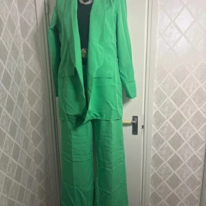 Grön kavaj och byxa set - Snyggt grönt set med kavaj och vida byxor. Kavajen har en klassisk design med långa ärmar och fickdetaljer. Perfekt för en stilren look.