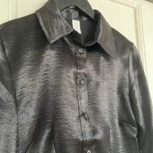 Svart skjorta från C.W. - Snygg svart skjorta från C.W. i glansigt material. Skjortan har långa ärmar och knappar framtill. Perfekt för en stilren look.