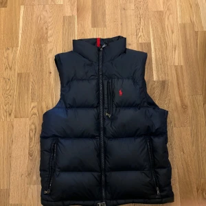 Svart dunväst från Polo Ralph Lauren - Snygg svart dunväst från Polo Ralph Lauren med röd broderad logga på bröstet. Västen har en dragkedja framtill och är perfekt för kyligare dagar. Den har en sportig look och är lätt att matcha med olika outfits.