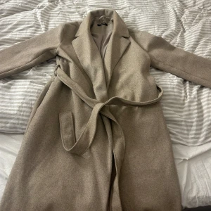 Beige kappa från Gina Tricot - Säljer en superfin beige kappa från Gina Tricot i storlek L. Den är i mycket bra skick och perfekt för höst och vår. Kappan har ett bälte i midjan och långa ärmar, vilket ger en elegant look. Materialet känns mjukt och bekvämt, perfekt för att hålla sig varm och stilren.