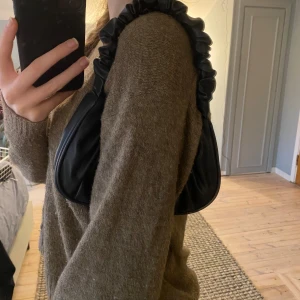 handväska - Säljer en superfin svart axelväska i syntetmaterial. Den har en unik rynkad rem som ger en cool look. Perfekt för både vardag och fest! Köpt för 150kr säljer för 45kr. Den är i mycket bra skick och redo för nya äventyr❣️ 