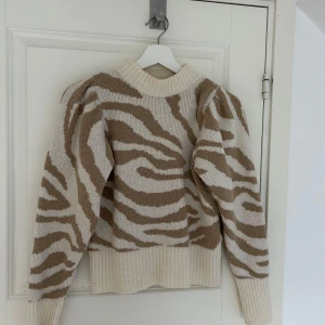 Beige och vit stickad tröja från Gina Tricot - Säljer en superfin stickad tröja från Gina Tricot i beige och vitt zebramönster. Den har långa ärmar och en bekväm ribbad kant vid halsen och ärmsluten. Perfekt för höst och vinter, både mysig och stilren! 🦓✨