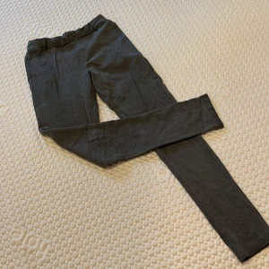 Grå byxor från Name It - Säljer ett par grå byxor från Name It i storlek 164. De ser ut som chinos men är mer som mjukisbyxor. De har fickor framtill och  hällor för bälte men även elastiskt dragskåp på insidan. Det som ser ut som en gylf är bara en detalj, inget som går att öppna.  Samma med bakfickorna. Byxorna är i använt skick och tyget ser lite nopprigt ut. 