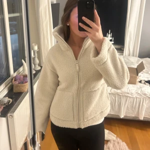 Beige teddyjacka med dragkedja - Mysig beige teddyjacka med dragkedja och hög krage. Perfekt för kyliga dagar med sin fluffiga och varma design. Jackan har långa ärmar och praktiska fickor framtill.