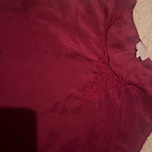 Vinröd ärmlös topp - Snygg vinröd ärmlös topp i ett mjukt och lätt material. Perfekt för sommardagar eller som ett lager under en kavaj. Den har en enkel design med en subtil detalj vid halsringningen som ger en extra touch. Passar både till vardags och festliga tillfällen.