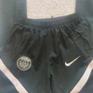 Svarta shorts från Nike med PSG-logga - Säljer ett par svarta byxor från Nike med Paris Saint-Germain-logga. De har en elastisk midja och är perfekta för träning eller vardagsbruk. Byxorna har en snygg detalj med rosa ränder längs sidorna och är gjorda i ett bekvämt material. Har aldrig använt dom!💕