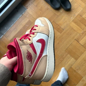 Nike Air Jordans i beige och röd - Säljer ett par Nike Air Jordans i beige och röd färg. Skorna har en snygg design med röd snörning och det klassiska Air Jordan-logotypen på sidan. De är i bra skick och kommer med originalkartong. Perfekta för både vardag och speciella tillfällen!